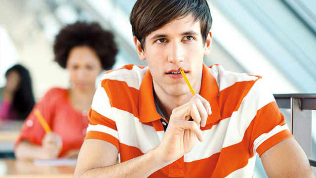 Ein junger Mann sitzt an einem Tisch, er hält einen Bleistift an seinen Mund. 